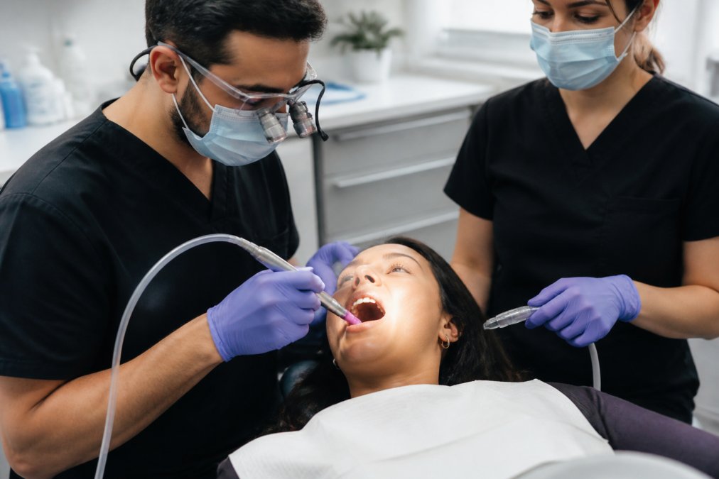 A dental professional in gloves and a mask holding a handheld laser instrument near a patient's open mouth in a bright, modern treatment room, with a dental assistant nearby