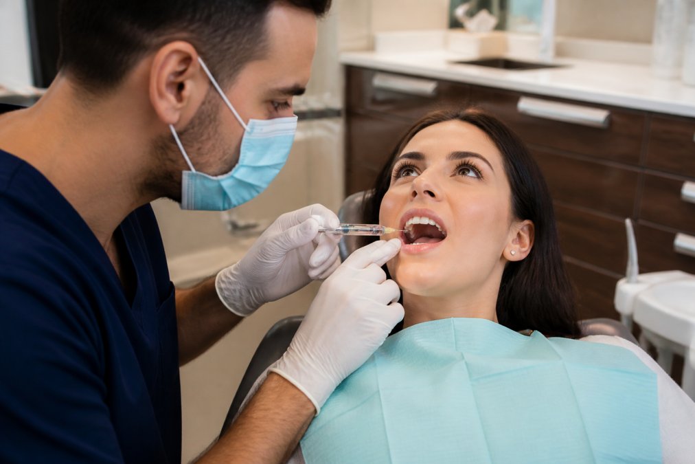 Professional dentist administering local anesthesia injection with calm patient in treatment chair, modern clinical setting with soft lighting and monitoring equipment visible
